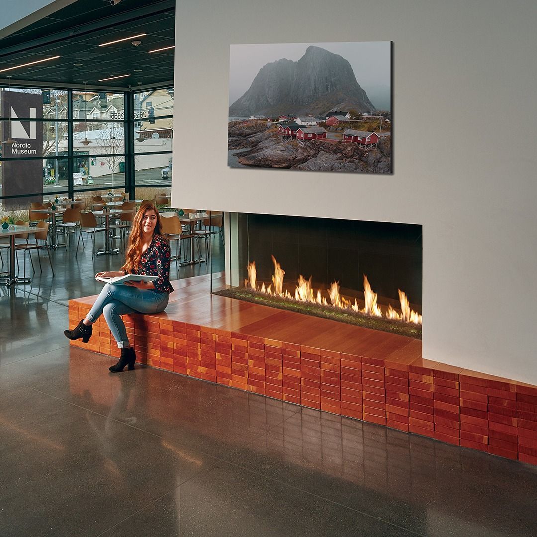 Woman sitting near a fireplace with flames, looking at a book. Mountain scene picture above.
