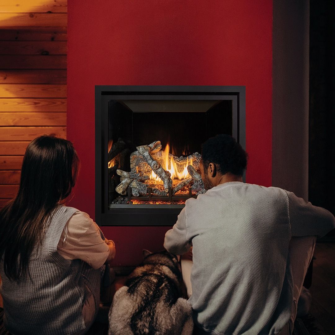 Couple and dog sitting by a burning fireplace with red wall.