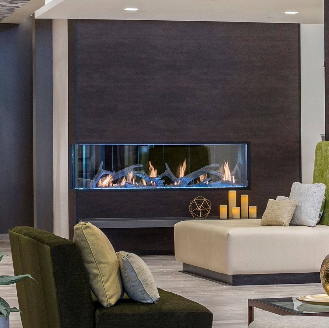 Cozy living room with a fireplace, dark wood paneling, and a beige ottoman and green chairs.