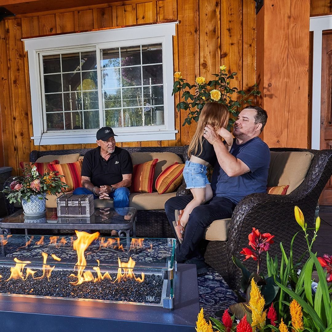 Family enjoying an outdoor patio. A man holds a child as another man sits on the couch near a fire pit.