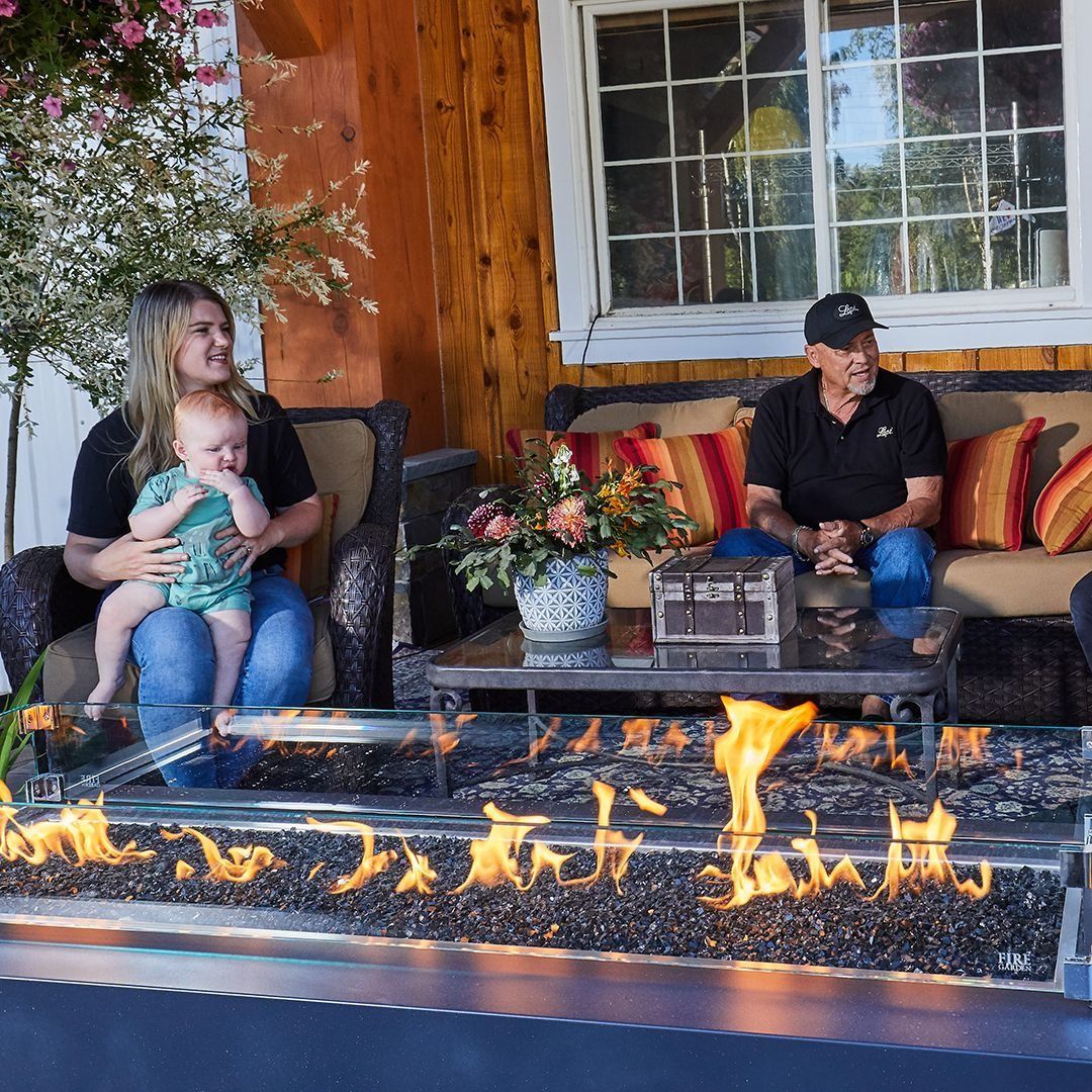 Family sits on a patio with a gas fire pit. A woman holds a baby, a man sits on a couch. Sunlit outdoor setting.