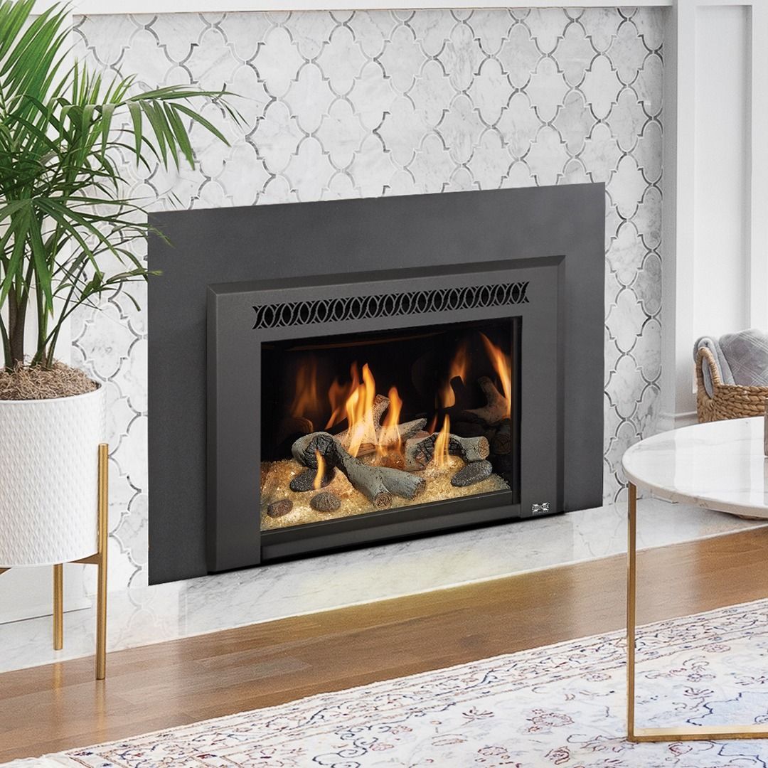 Fireplace with black frame, burning logs, on white tile wall. Plant and table in the room.