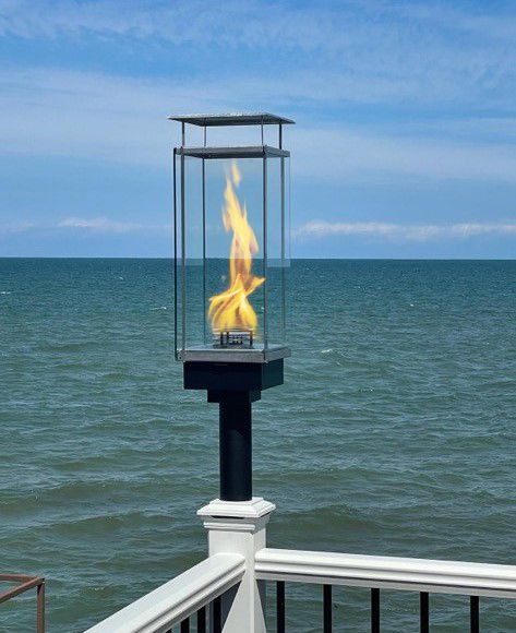 Flaming gas lamp on a black post, glass enclosure. Ocean background, blue sky.