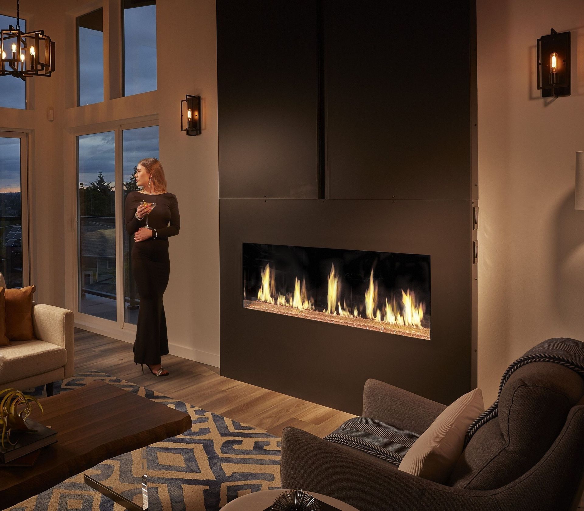 Woman in black dress by fireplace with flames; modern living room at dusk.