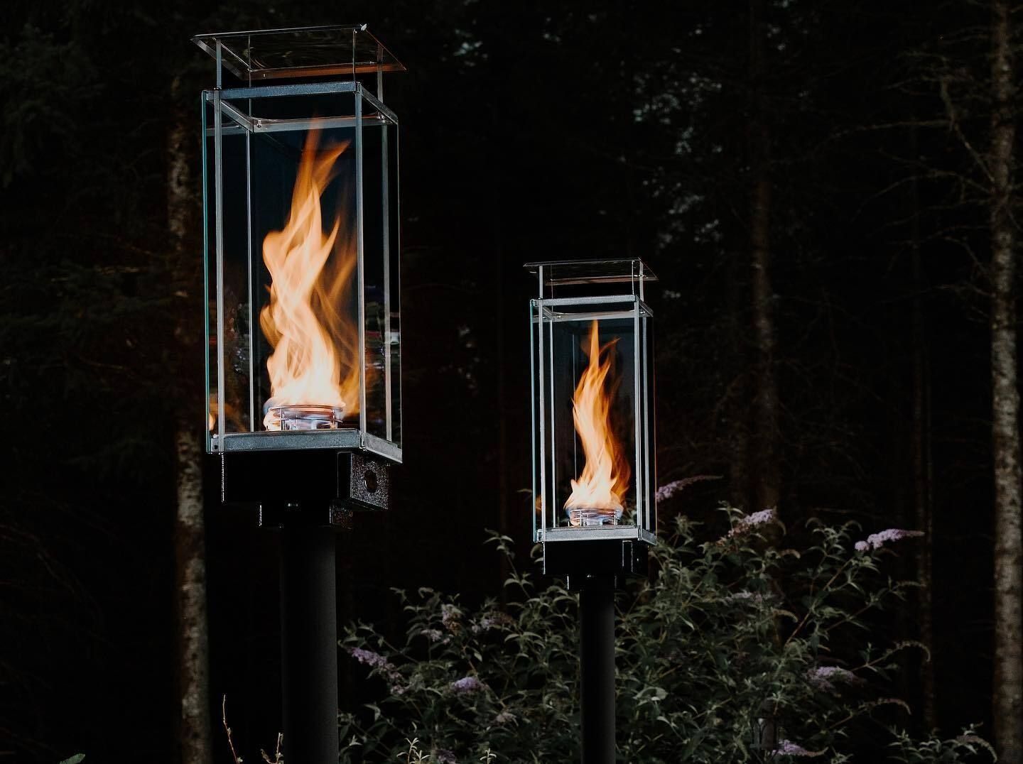 Two lit, caged flames atop posts in a dark forest.