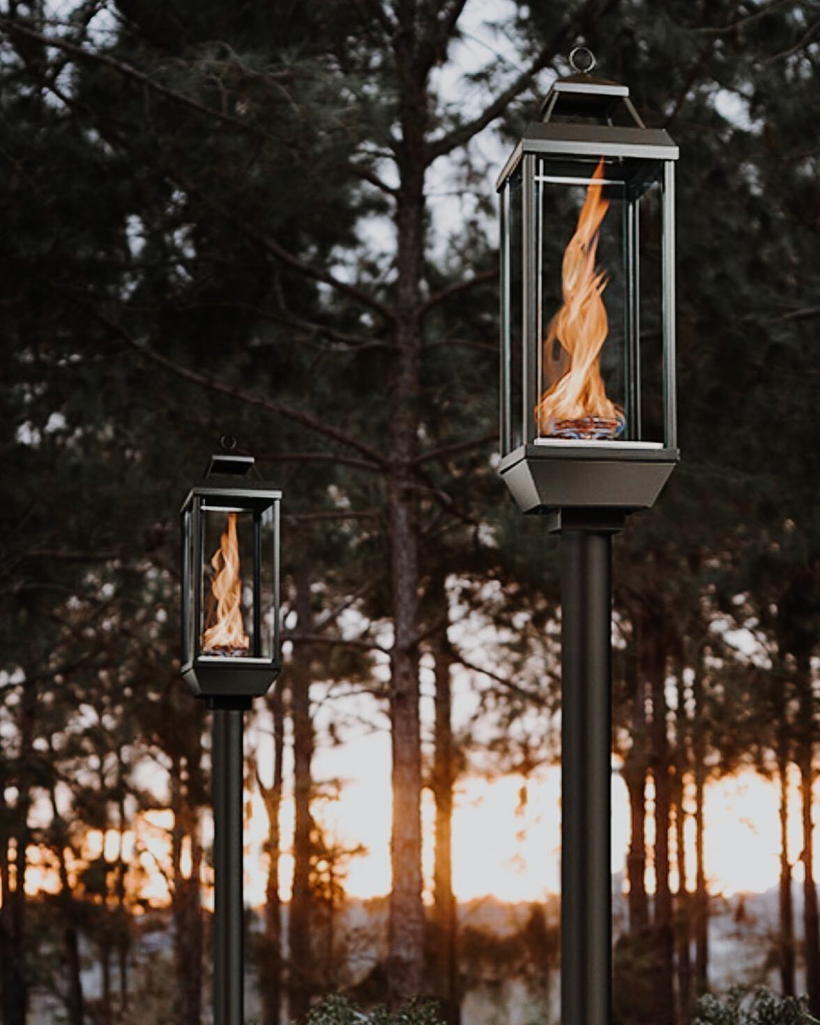 Two outdoor gas lanterns with flames, set against a blurred forest at dusk.