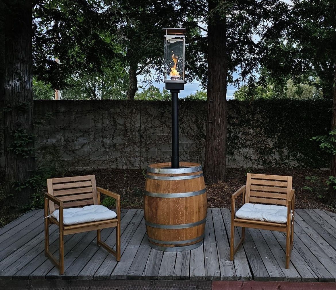 Wooden chairs face a fire pit on a deck. A barrel base supports the black pole and glass-enclosed flames.