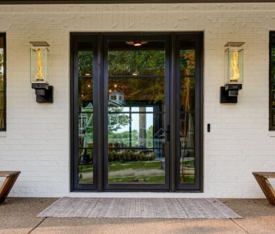 Front door with black frame, glass panels, flanked by sconces on white brick wall.