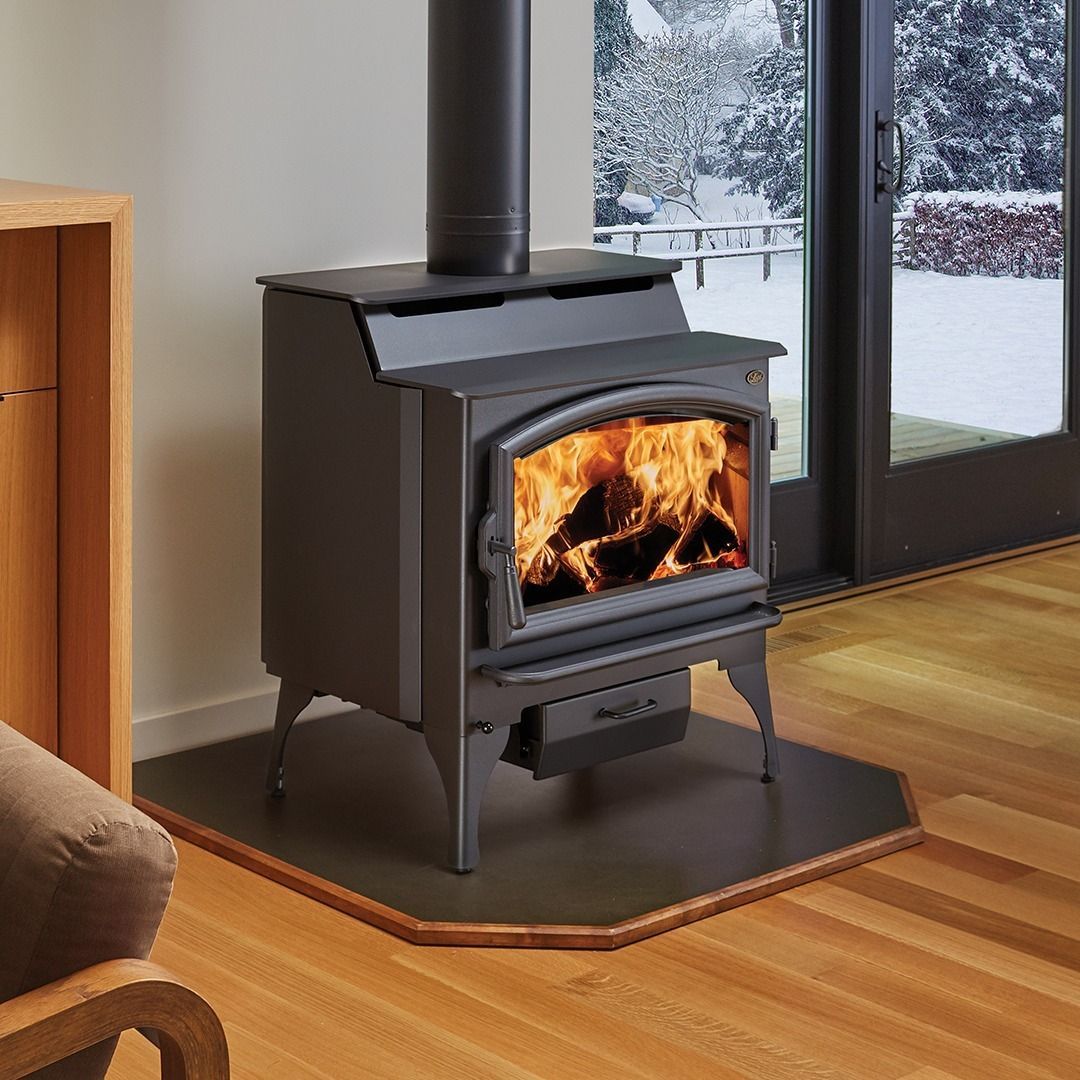 A black wood-burning stove with a lit fire, sits on a stone hearth in a room with a window.