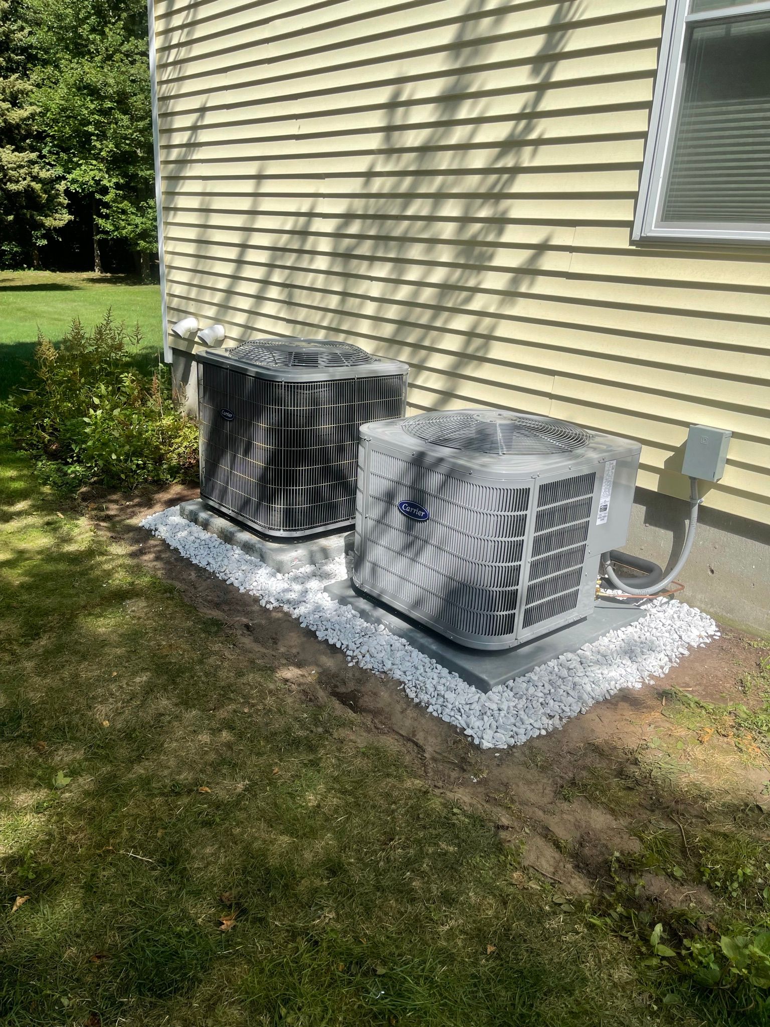 Two air conditioning units on a gravel bed next to a yellow house with a window.
