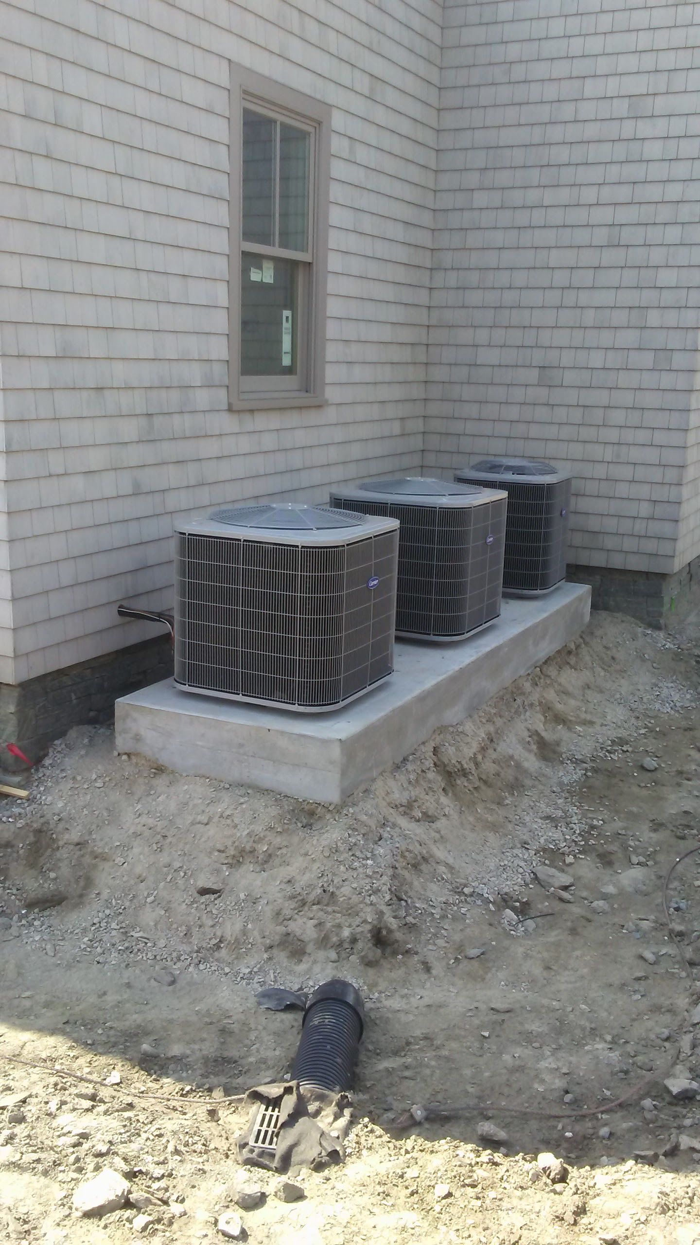 Three air conditioning units on a concrete pad outside a building with weathered siding and a window.