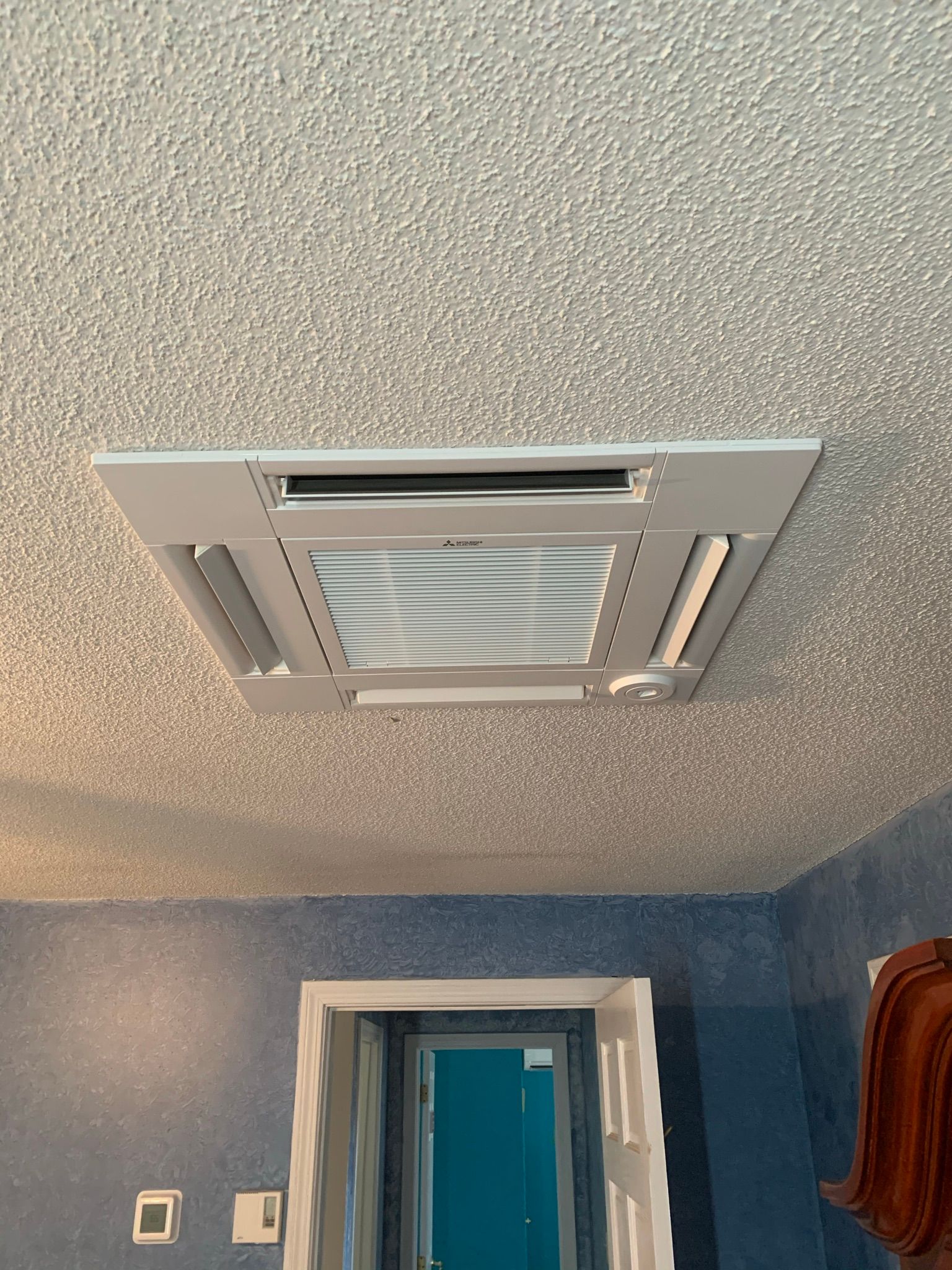 Ceiling-mounted air conditioner in a room with textured ceiling, blue walls, and an open doorway.
