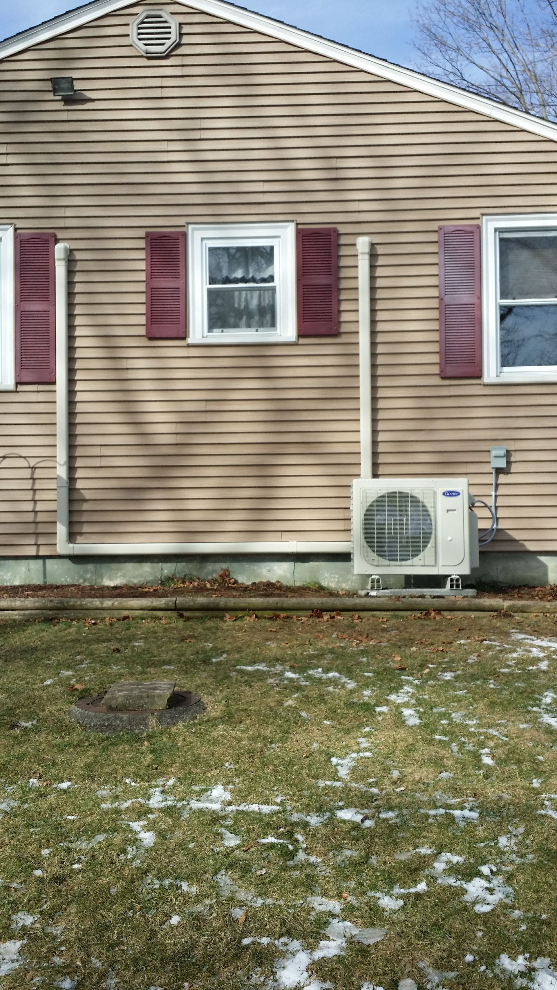 Tan house exterior with red shutters. Air conditioning unit on the ground, beige. Partially snow-covered lawn.