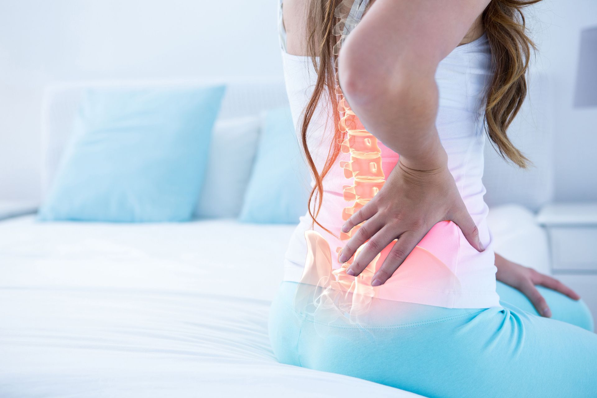 A woman is sitting on a bed holding her back in pain.