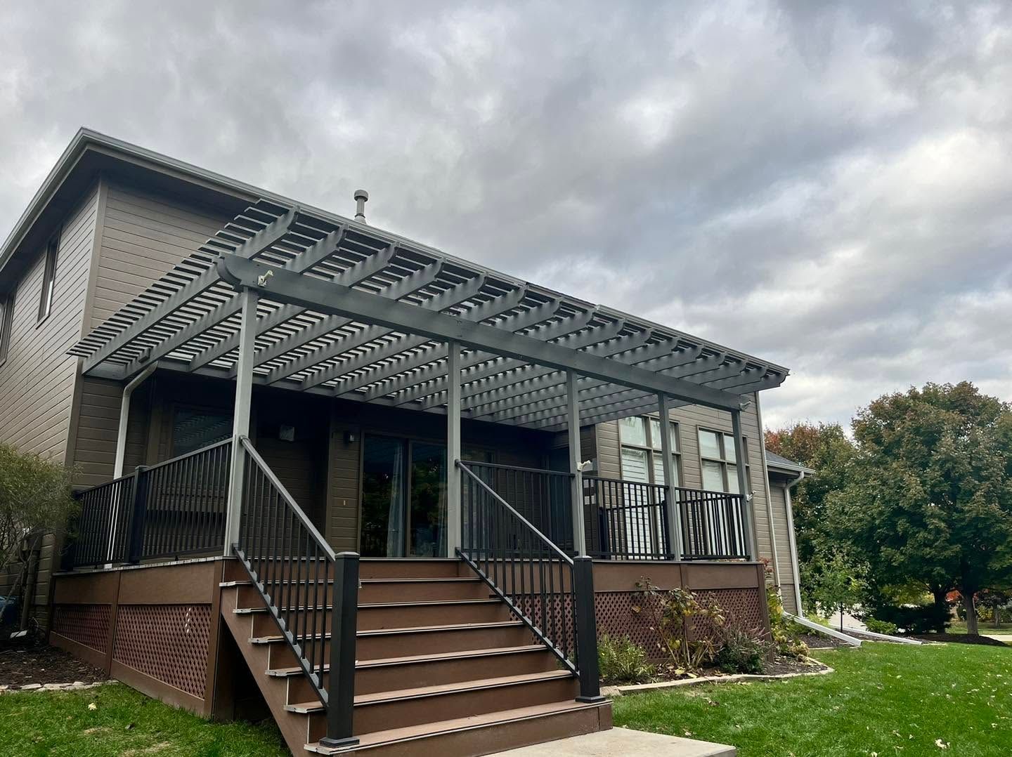 Front porch with staircase, black railing, and gray pergola roof on a brick house under cloudy skies
