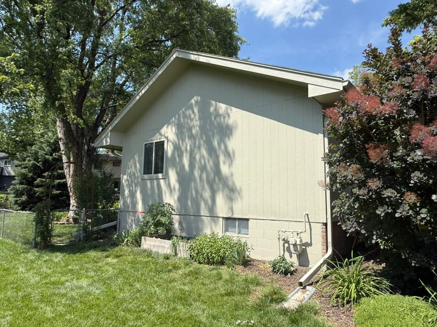 Side view of a beige house with trees, bushes, and a grassy yard under a sunny sky