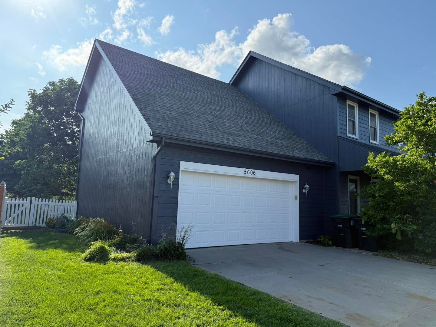 Dark gray suburban house with white garage door and driveway in front, under a partly cloudy sky