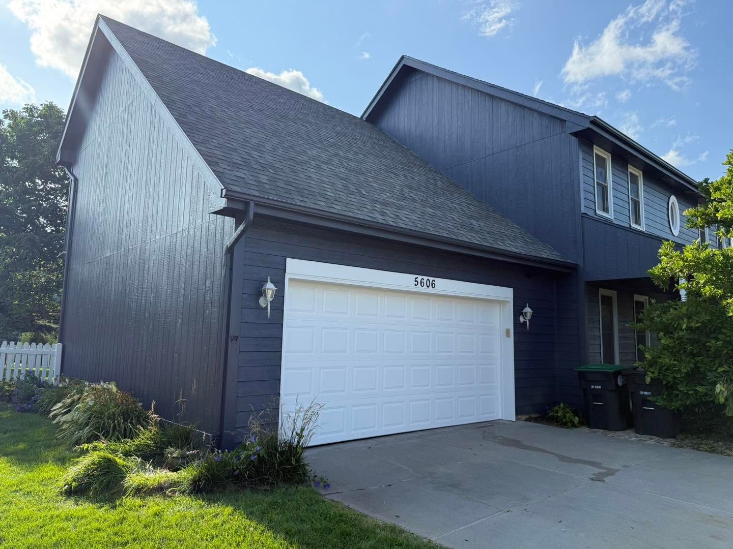 Dark blue two-story house with a white garage door and driveway under a sunny sky