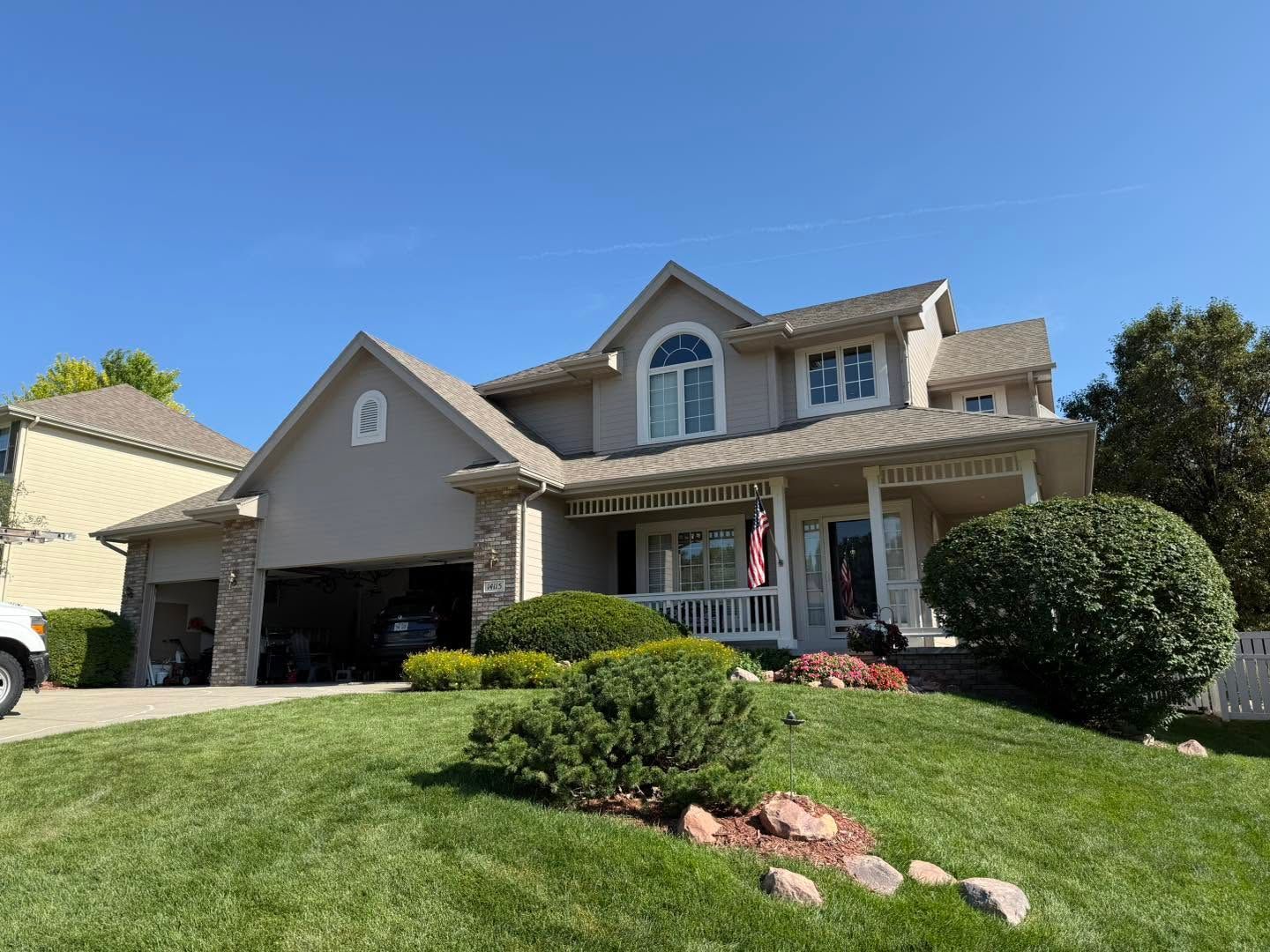 Suburban two-story house with beige siding, front porch, manicured lawn, and blue sky