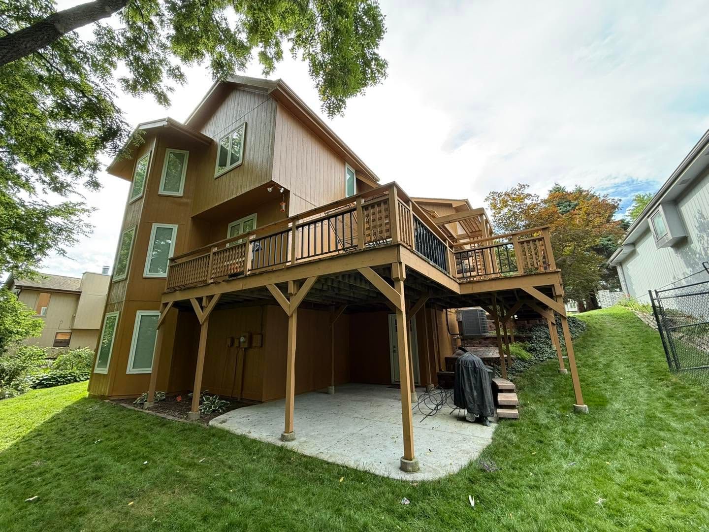 Two-story brown house with wooden deck, stairs, and grassy yard under trees