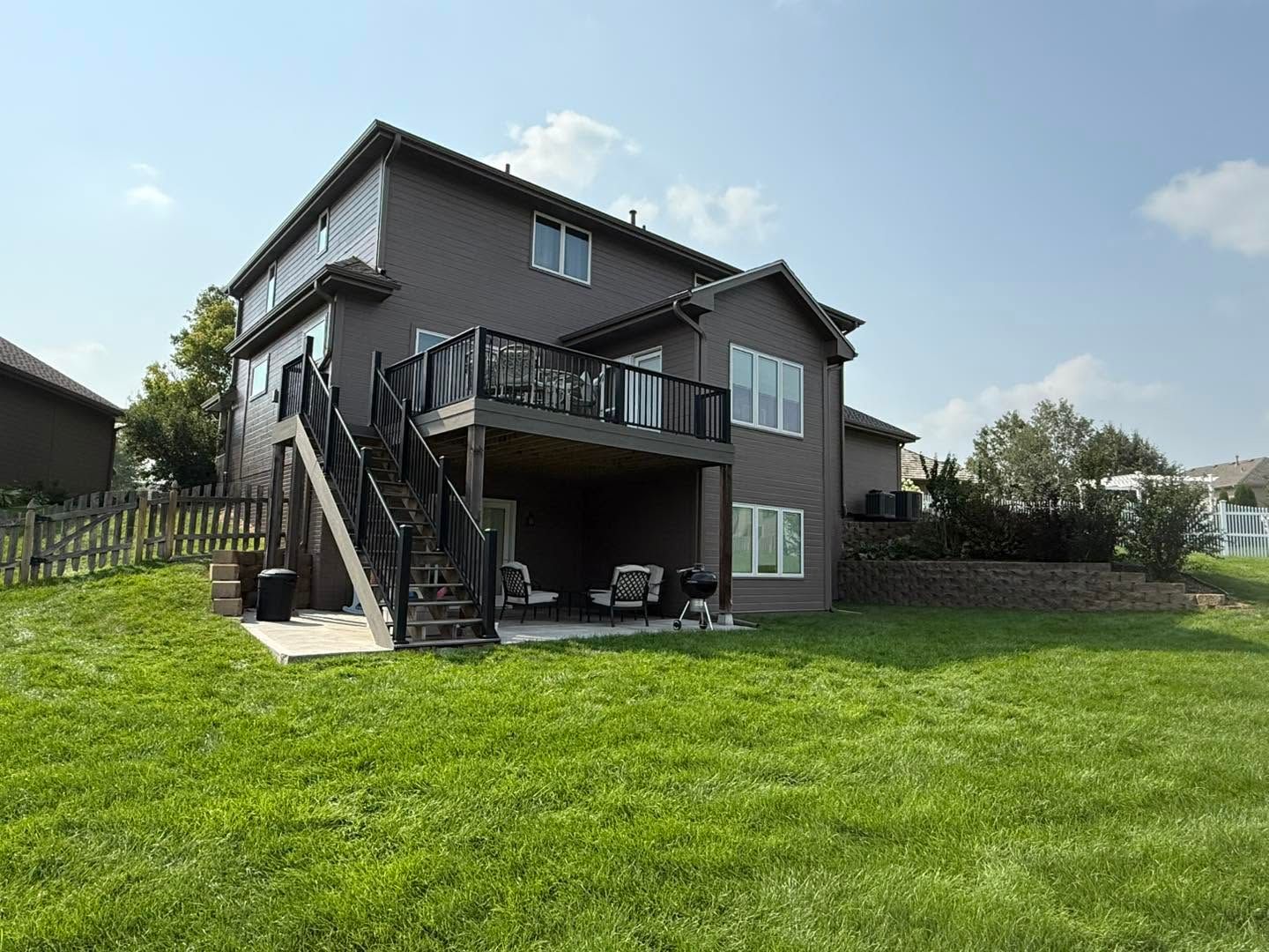 Two-story house with a wooden deck and stairs, overlooking a large green lawn under a clear sky