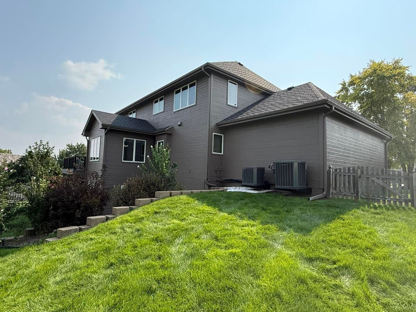 Gray two-story house with a sloped green lawn and blue sky in the background