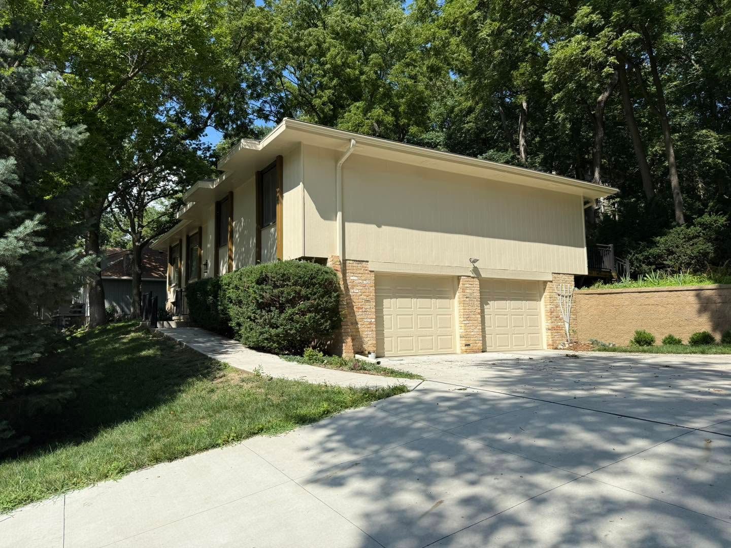 Beige two-car garage beside a wooded driveway with a brick retaining wall.