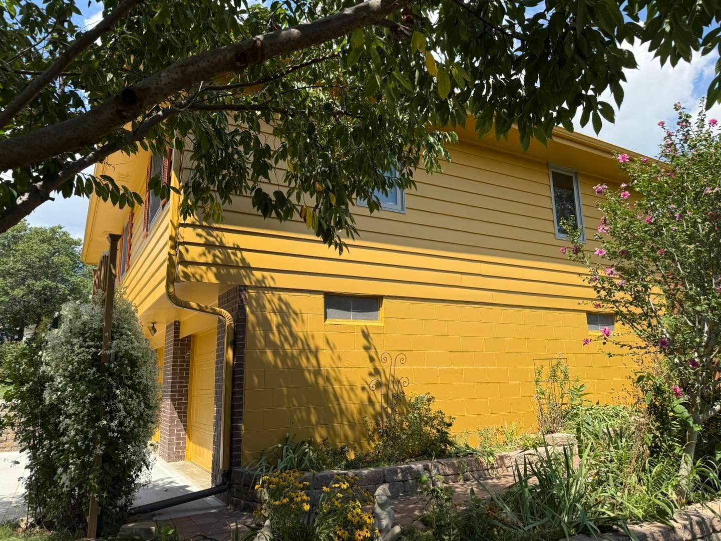 Yellow two-story house with a front entry, trees overhead, and garden plants along the wall