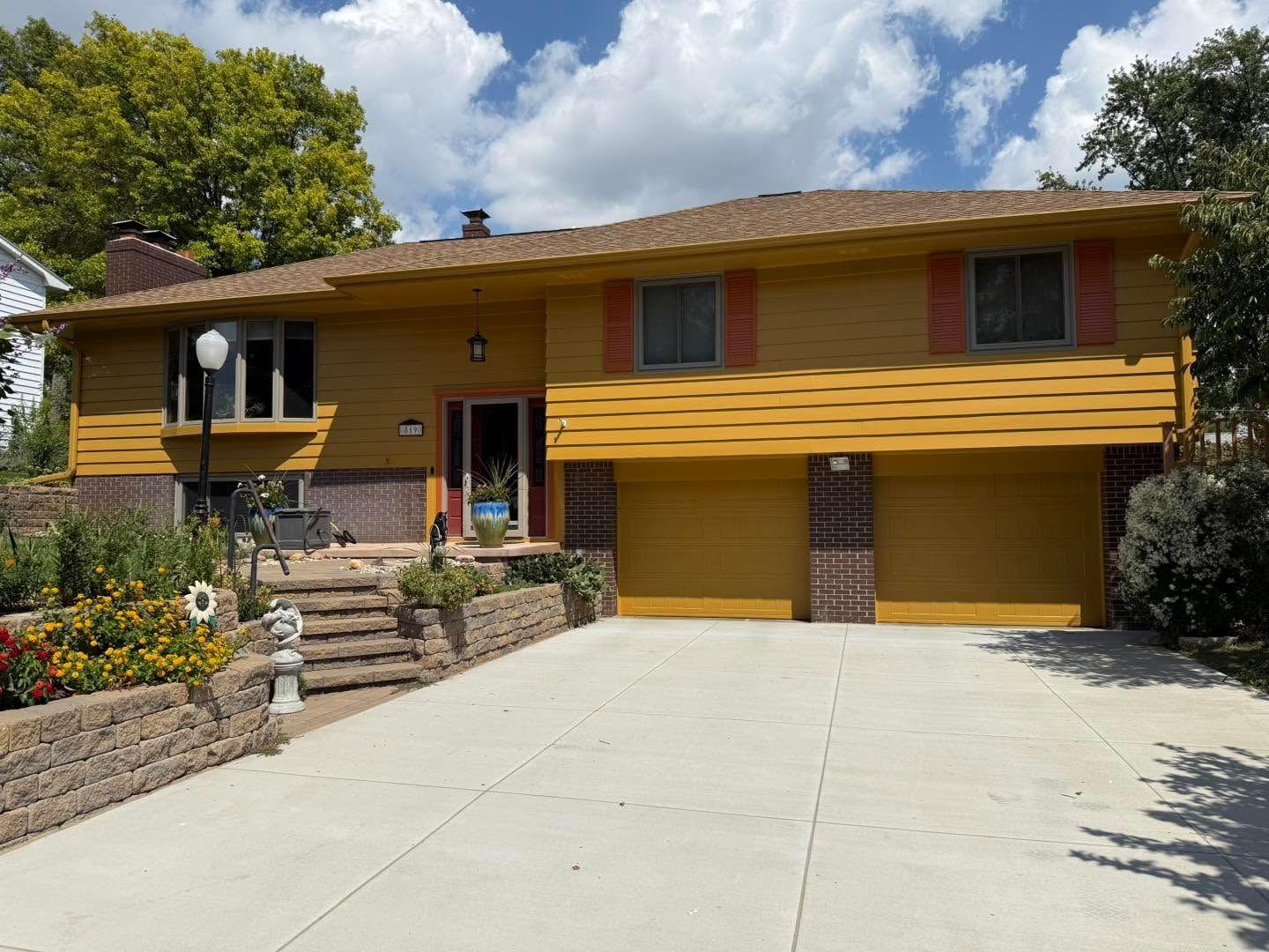 Yellow two-story house with red shutters and a wide driveway under a partly cloudy sky
