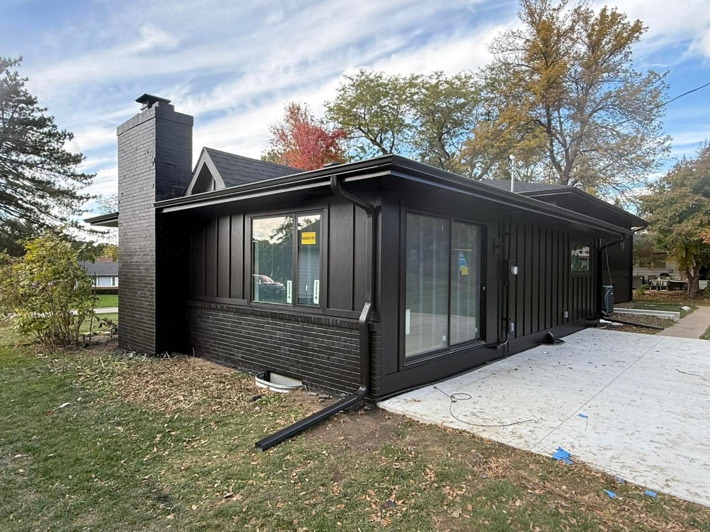 Modern black house exterior with large windows, chimney, and driveway beside autumn trees