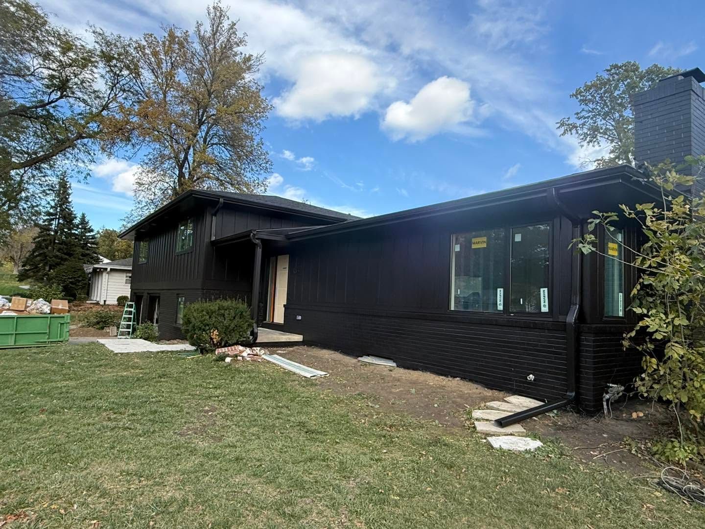 Single-story dark house with large windows, grassy yard, and a tree under a partly cloudy sky