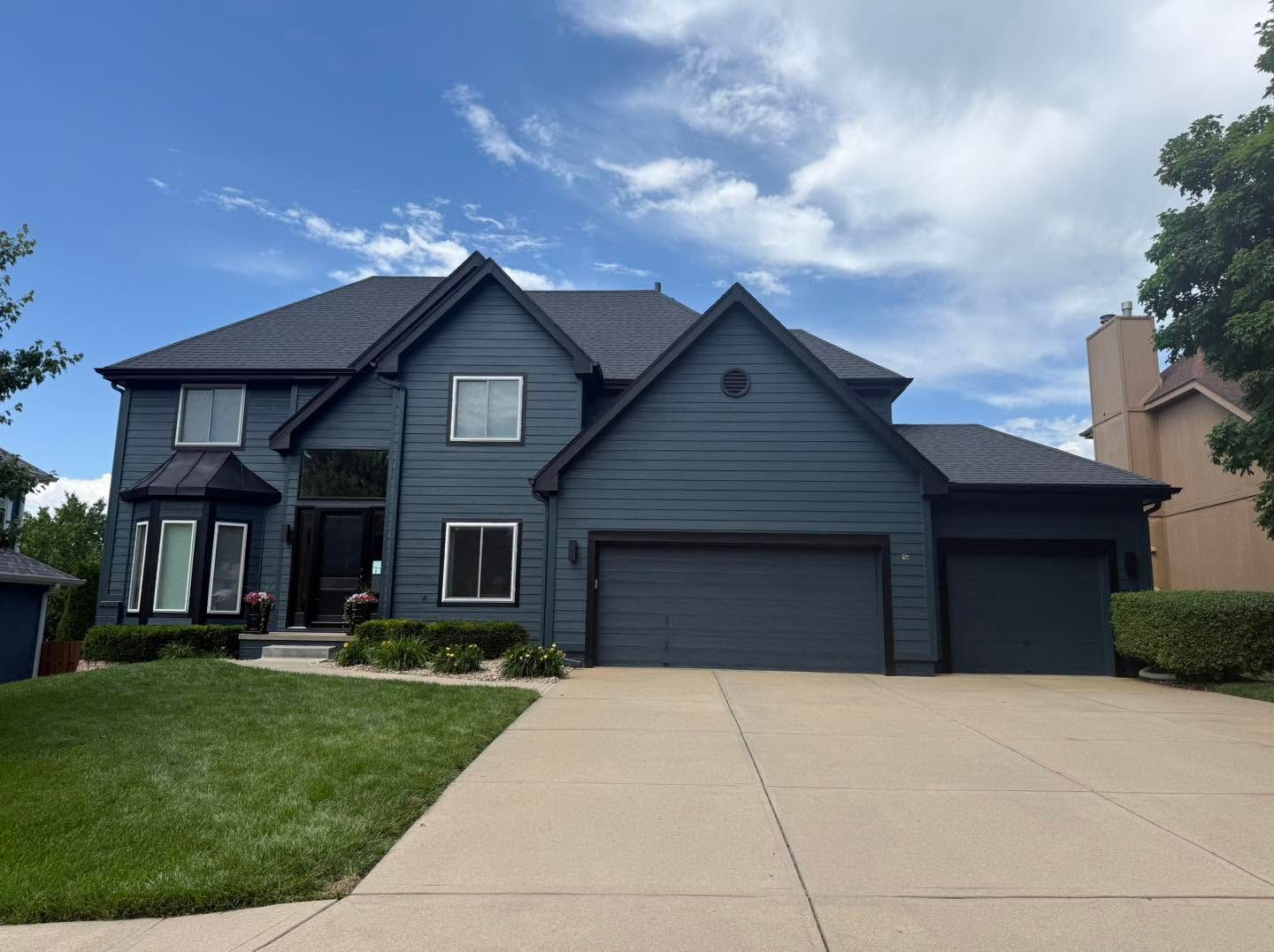 Gray two-story suburban house with a three-car garage and a large driveway under a partly cloudy sky