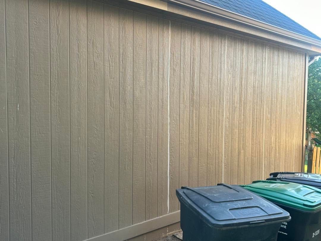 Wooden house siding with blue roof edge and two trash bins along the side yard.