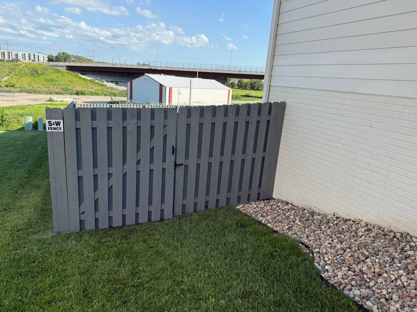 Gray slatted fence beside a white building on a grassy yard with gravel edging.