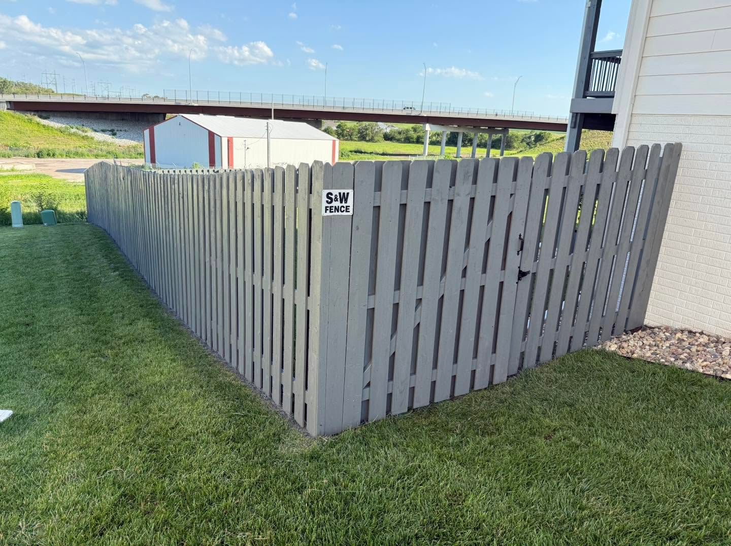 Gray wooden fence around a grassy yard beside a house under a blue sky