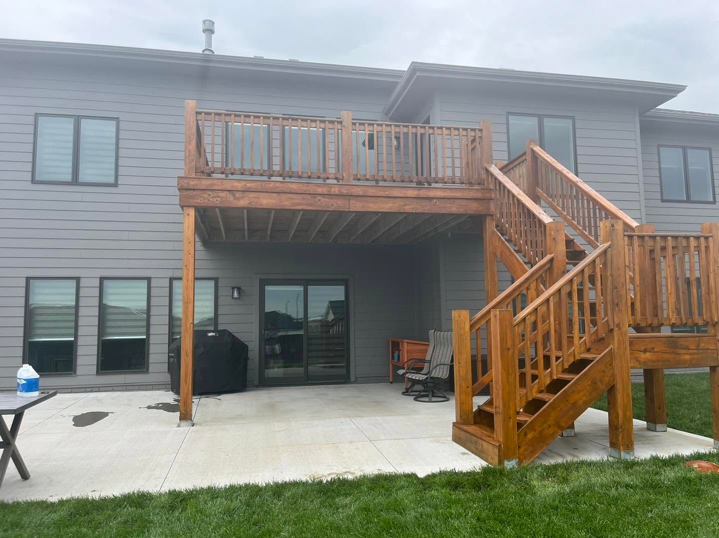 Two-story gray house with wooden deck and stairs overlooking a concrete patio and green lawn.
