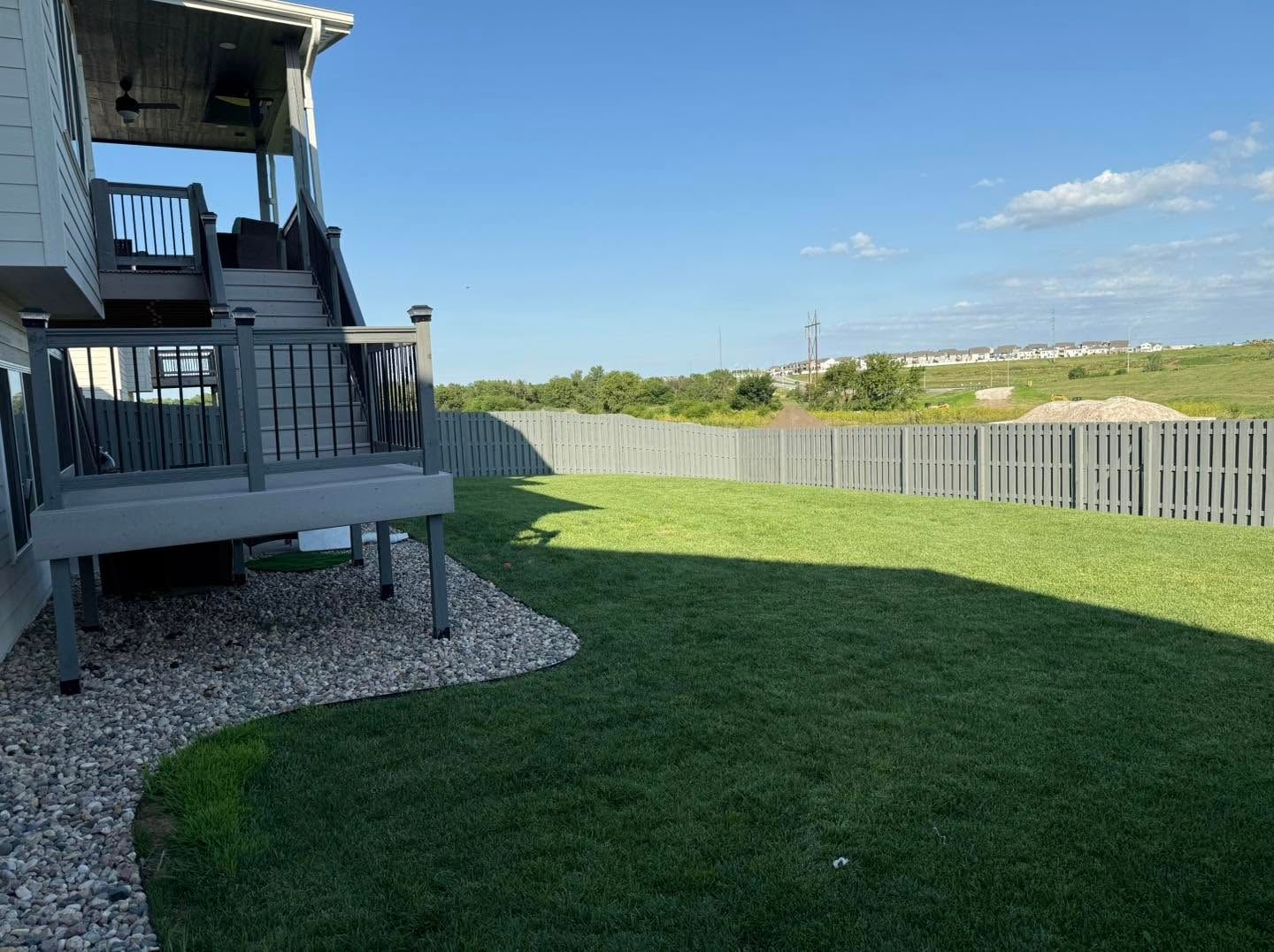 Backyard with green lawn, gray fence, and a deck beside a house under a blue sky