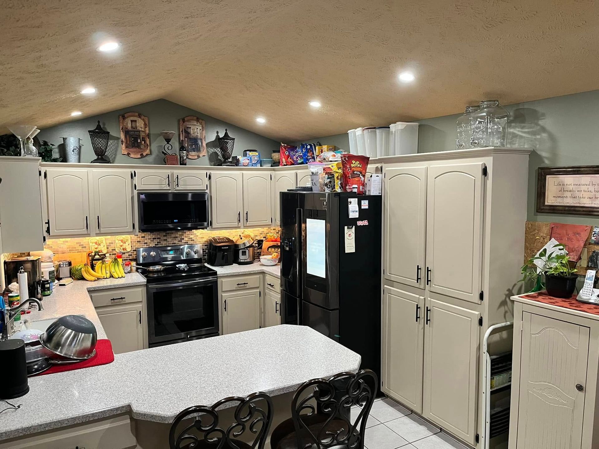 Bright kitchen with white cabinets, black stove, double fridge, and a granite island under recessed lights