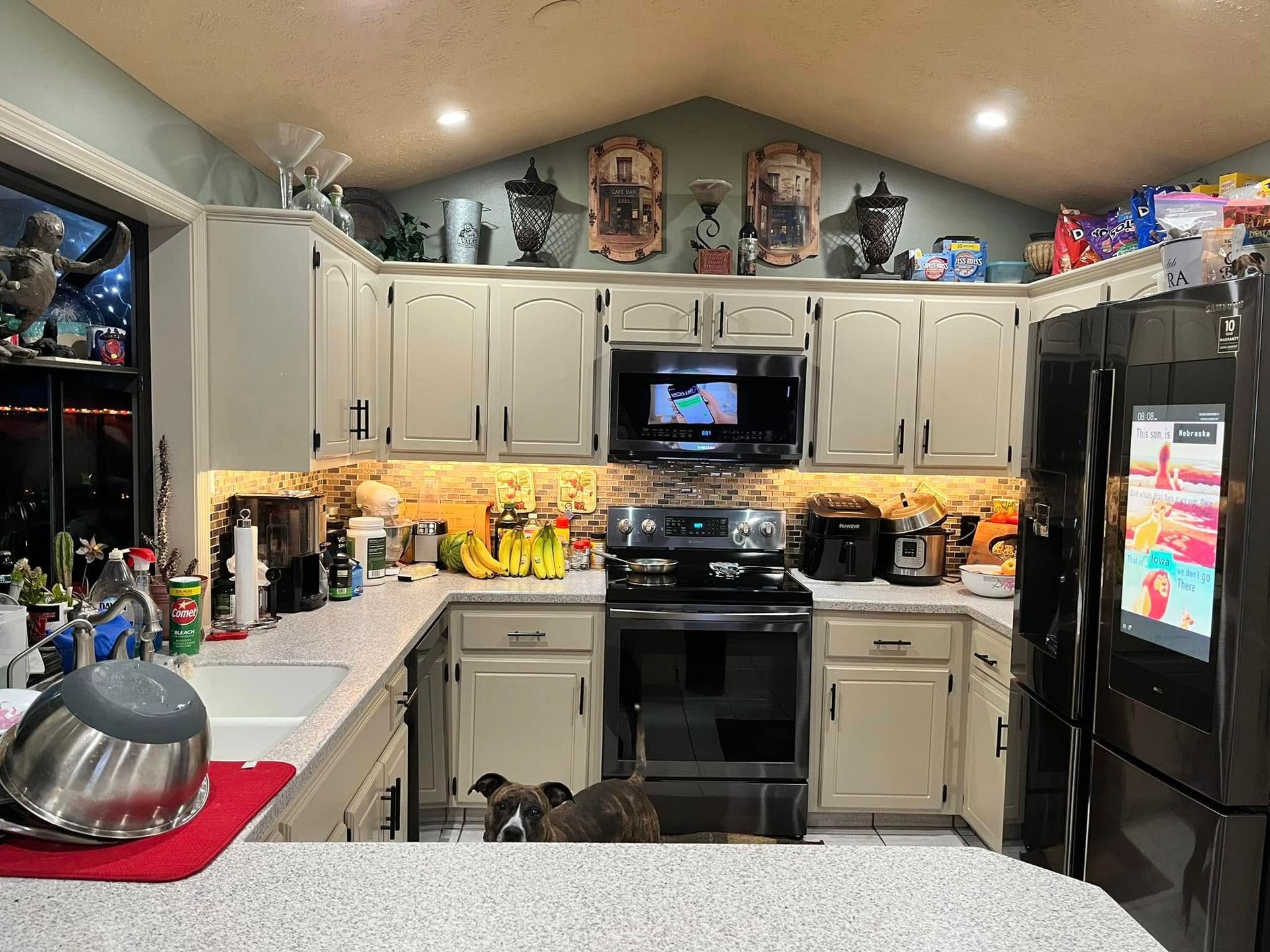Cluttered kitchen with white cabinets, black appliances, red dish towel, and decorative items on counters and above cabinets