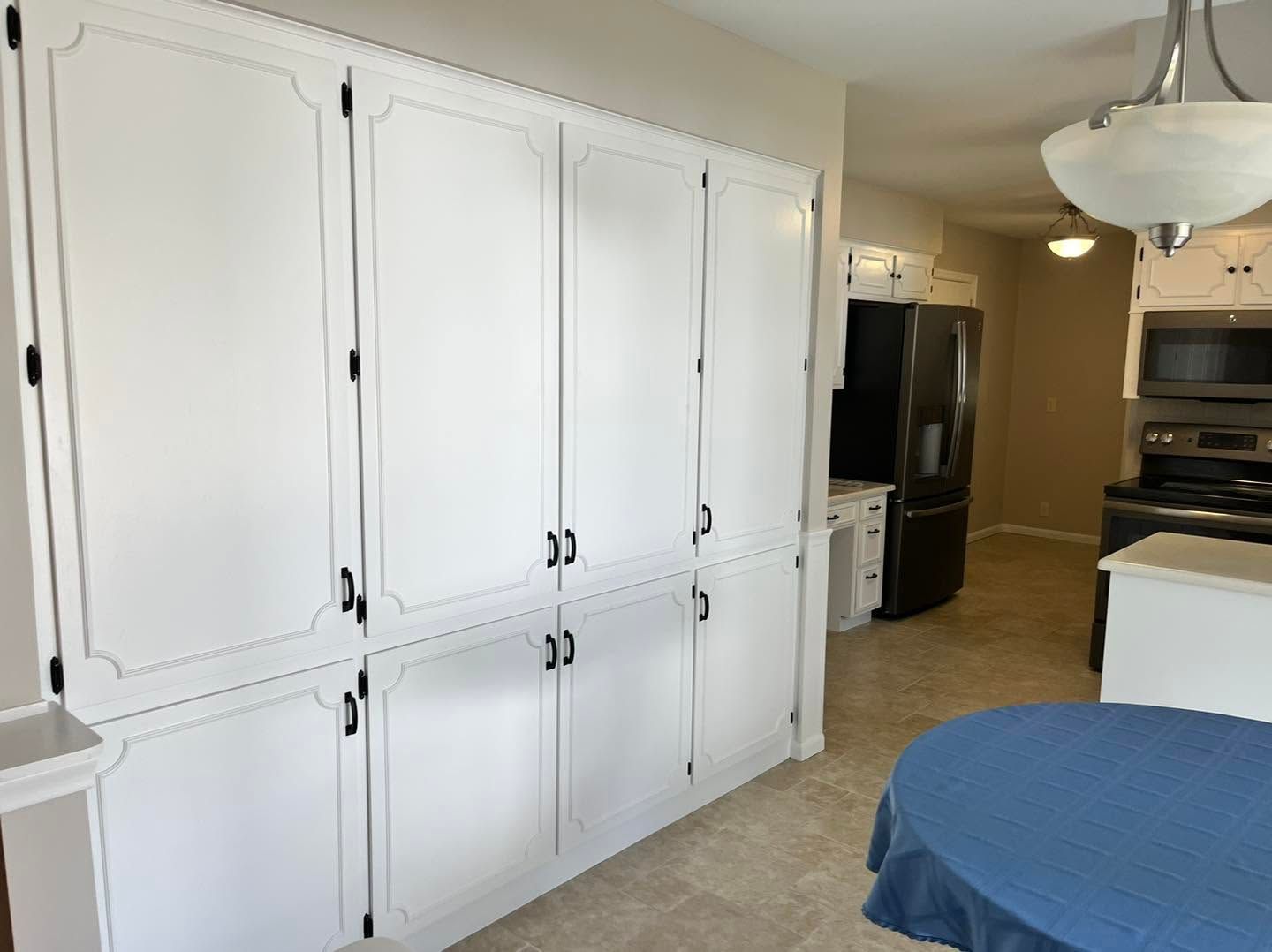 White kitchen cabinets beside a bedroom area with a bed, ceiling lights, and black refrigerator in the background