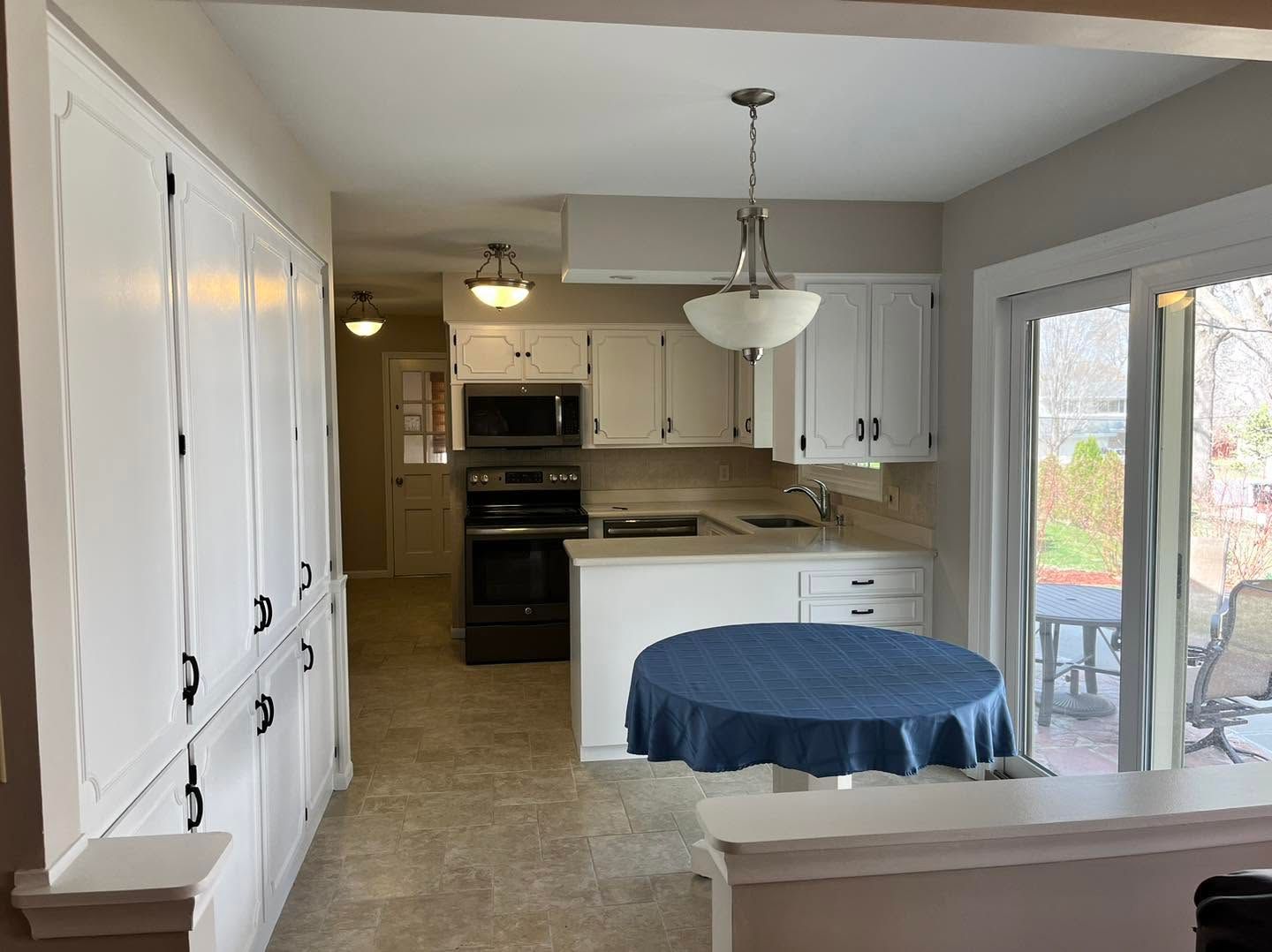 Bright kitchen with white cabinets, tiled floor, and a round table covered in blue cloth by sliding glass doors.