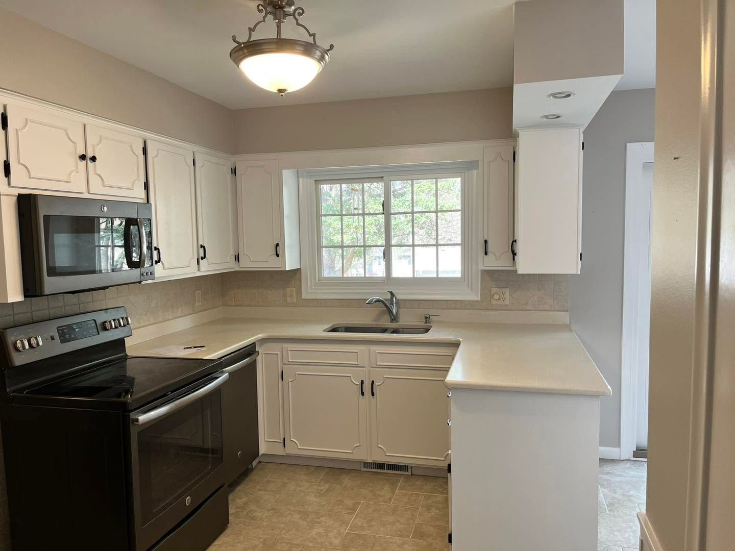 Small kitchen with white cabinets, black stove, beige countertops, and a window over the sink.