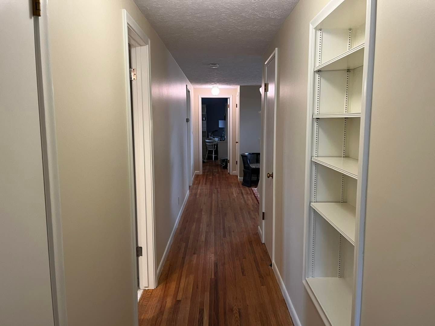 Narrow hallway with hardwood floors, beige walls, and white built-in shelves on the right.