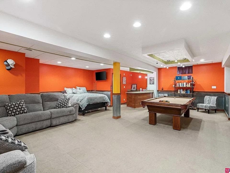 Basement rec room with orange walls, gray sectional sofa, pool table, beds, and shelving