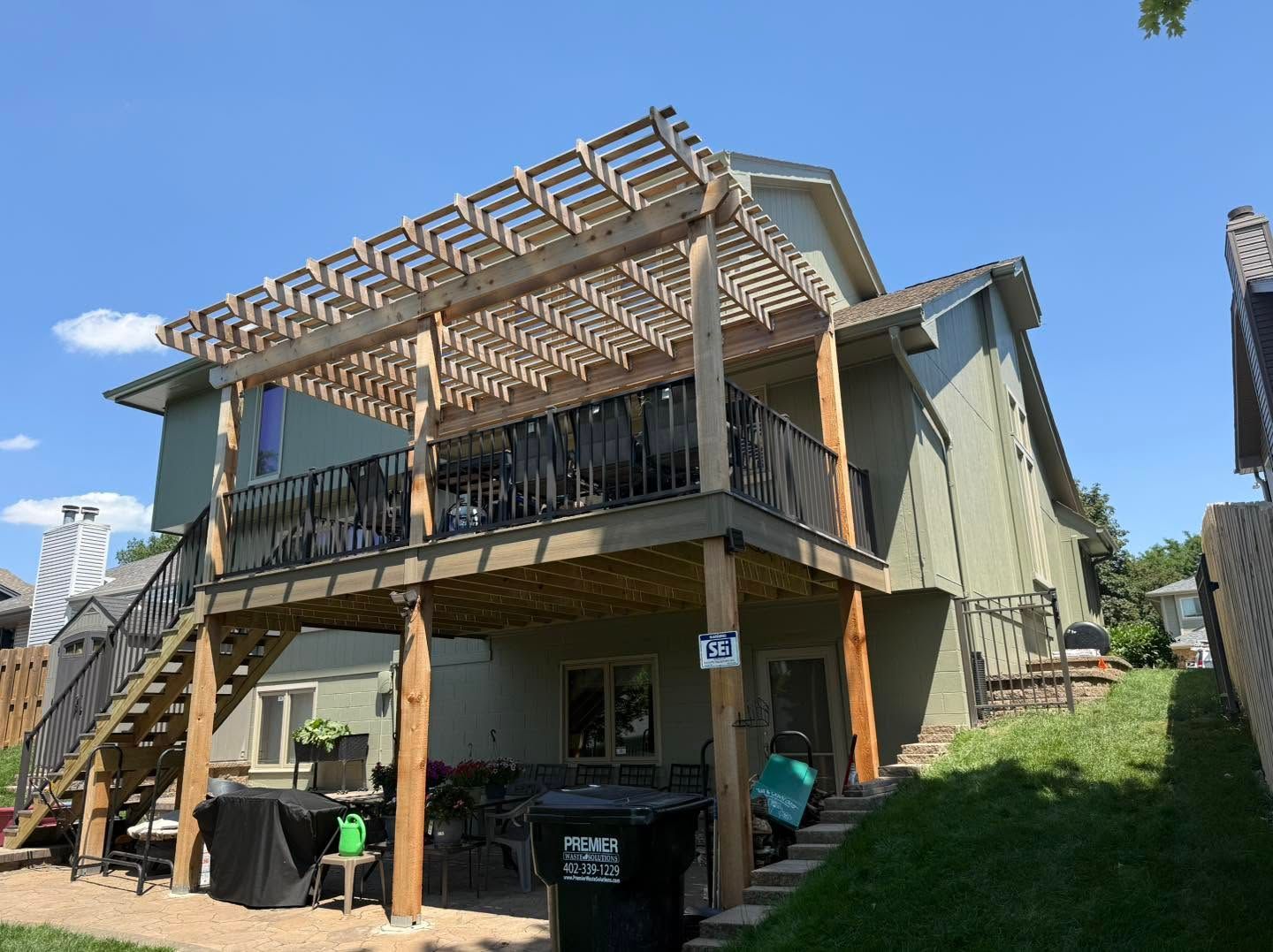 Backyard deck with pergola over a two-story house, stairs at left, and trash bins below