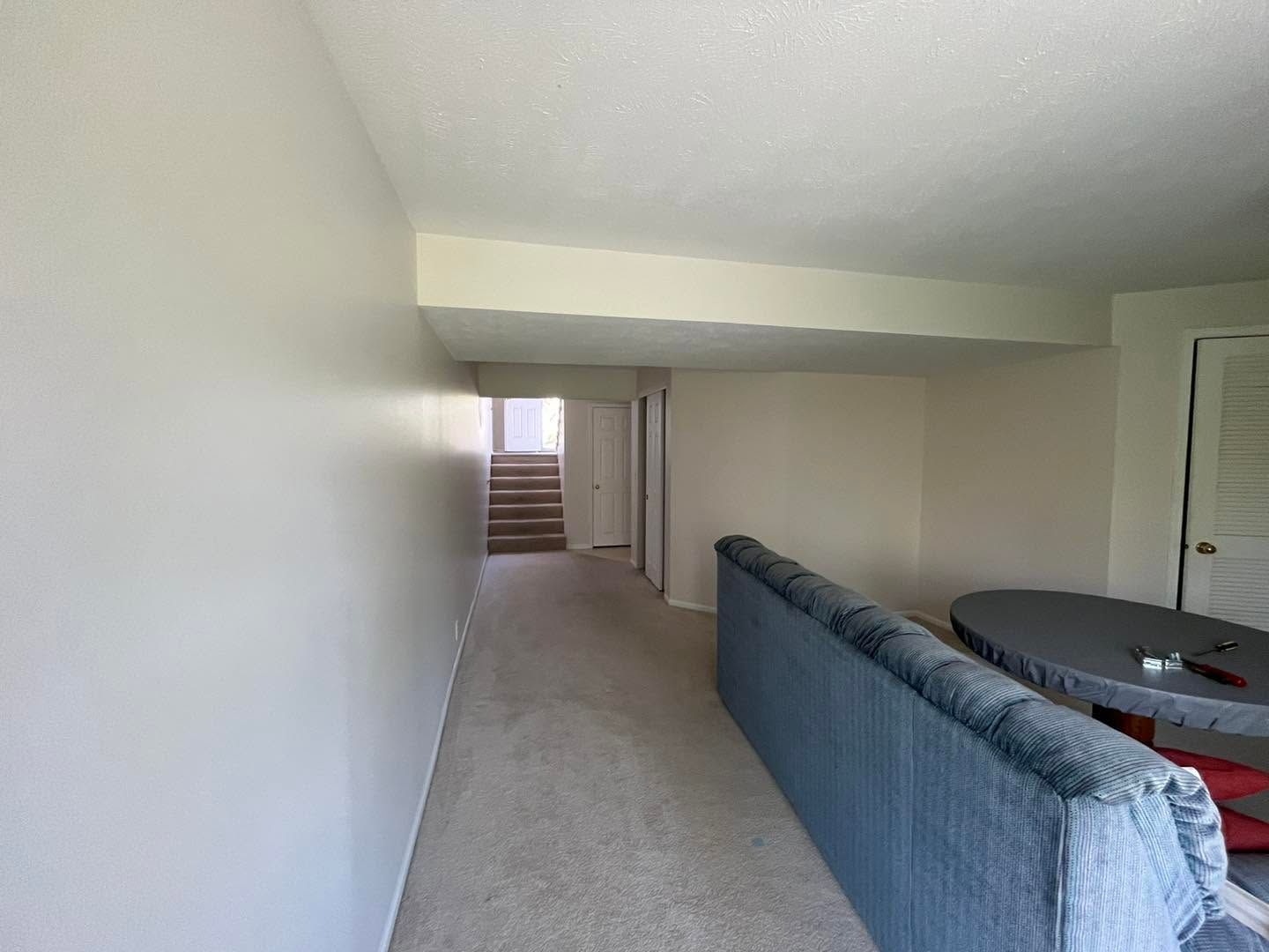 Long carpeted hallway leading to stairs, with a blue sofa and dining table in a beige room.