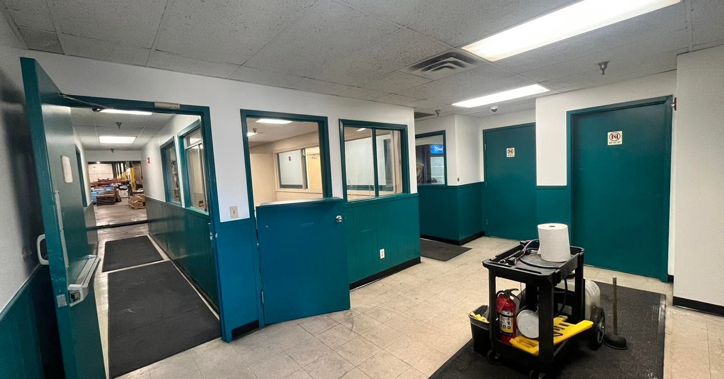 Empty office hallway with teal doors and partitions, lit by fluorescent lights, with a small cart in the foreground