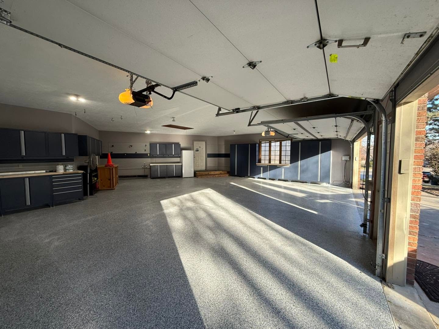 Empty garage with gray floor, overhead lighting, and sunlight streaming through the open door.