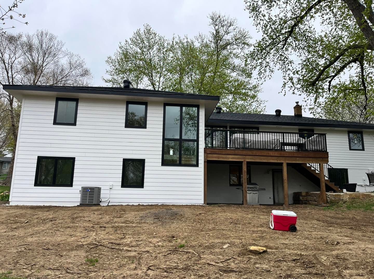 White two-story house with black trim, deck, and stairs in a wooded yard
