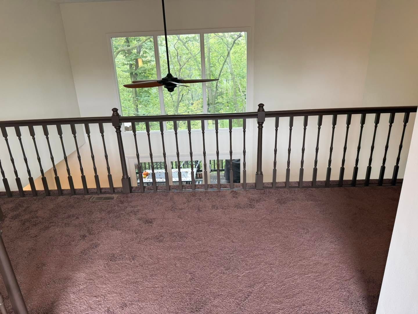 Empty upstairs landing with carpet, dark railing, ceiling fan, and a large window overlooking green trees